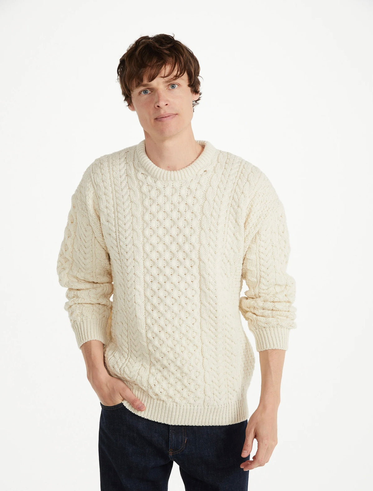 A man wearing a cream-colored cable-knit crewneck sweater and dark blue jeans, standing against a plain white studio background.
