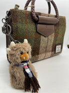 A close-up of a brown and green plaid tweed handbag with a braided brown handle, a leather tab, and a plush cow keychain accessory with a scarf.