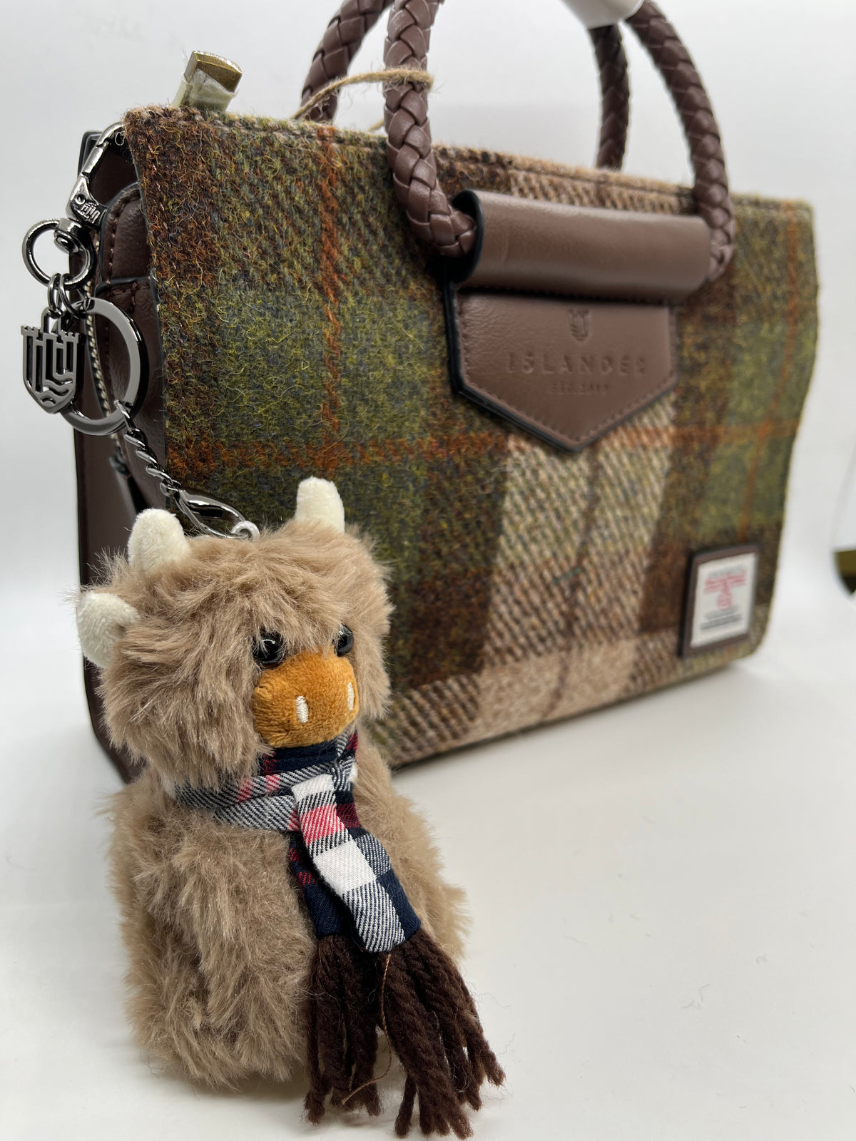 A close-up of a brown and green plaid tweed handbag with a braided brown handle, a leather tab, and a plush cow keychain accessory with a scarf.