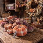 A collection of fall-themed decorative pumpkins in orange, purple, and beige plaid fabric, displayed on a rustic wooden table in a cozy autumn-themed shop setting.