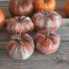 Four fabric pumpkins in a rustic autumn plaid pattern, arranged on a weathered wooden surface with real pumpkins in the background.