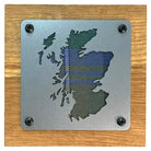 A square metal plaque featuring the silhouette of Scotland with a blue and green tartan plaid pattern, mounted on a square wooden board with four black screws in the corners.