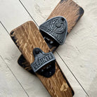 Two rustic wooden bottle opener plates with black metal 'SCOTLAND' and 'SLAINTE' branded wall-mounted bottle opener hardware, displayed on a light wood plank surface.