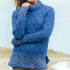 A woman wearing a blue cable-knit mock-neck sweater with white Celtic knot patterns on the cuffs and hem, photographed outdoors against a soft beach background.