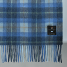 A close-up of a blue plaid wool blanket with fringe, featuring a black brand label that reads ISLANDER.
