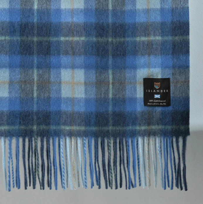 A close-up of a blue plaid wool blanket with fringe, featuring a black brand label that reads ISLANDER.
