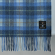 A close-up of a blue plaid wool blanket with fringe, featuring a black brand label that reads ISLANDER.