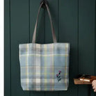 A plaid canvas tote bag with brown shoulder straps hangs on a dark green wooden wall. The bag features a light blue, yellow, and white plaid pattern with a small embroidered purple flower near the bottom right.