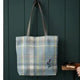 A plaid canvas tote bag with brown shoulder straps hangs on a dark green wooden wall. The bag features a light blue, yellow, and white plaid pattern with a small embroidered purple flower near the bottom right.