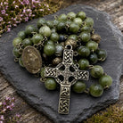 A green stone bead rosary with an antique bronze Celtic cross and medal, displayed on a dark slate stone with small pink flowers in the background.