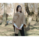 A woman wearing a camel-colored knit poncho draped over her shoulders, styled over a white turtleneck and black pants, standing in a park with blurred trees in the background.