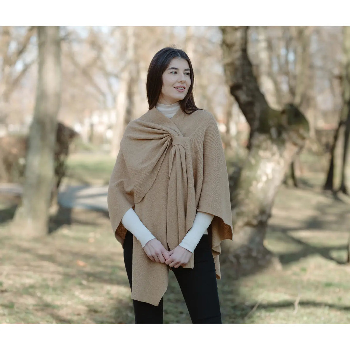 A woman wearing a camel-colored knit poncho draped over her shoulders, styled over a white turtleneck and black pants, standing in a park with blurred trees in the background.