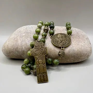 An olive green stone bead rosary with an antique bronze Celtic cross and medallion, displayed on a smooth light gray stone against a neutral gray background.
