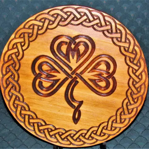 A round wooden decorative plaque featuring a Celtic knot border and a central shamrock design with heart-like details, shown on a dark textured background.