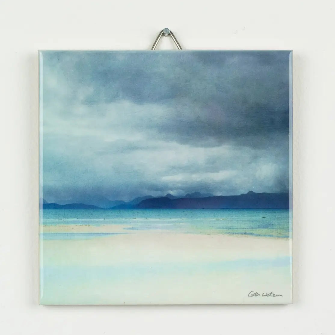 A square ceramic art tile wall art print depicting a misty blue and gray seascape with distant mountains and pale beach tones, hanging against a white wall with a metal hanger visible at the top.