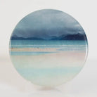A round ceramic coaster featuring a scenic landscape print of a beach with mountains, displayed on a white surface against a white background.