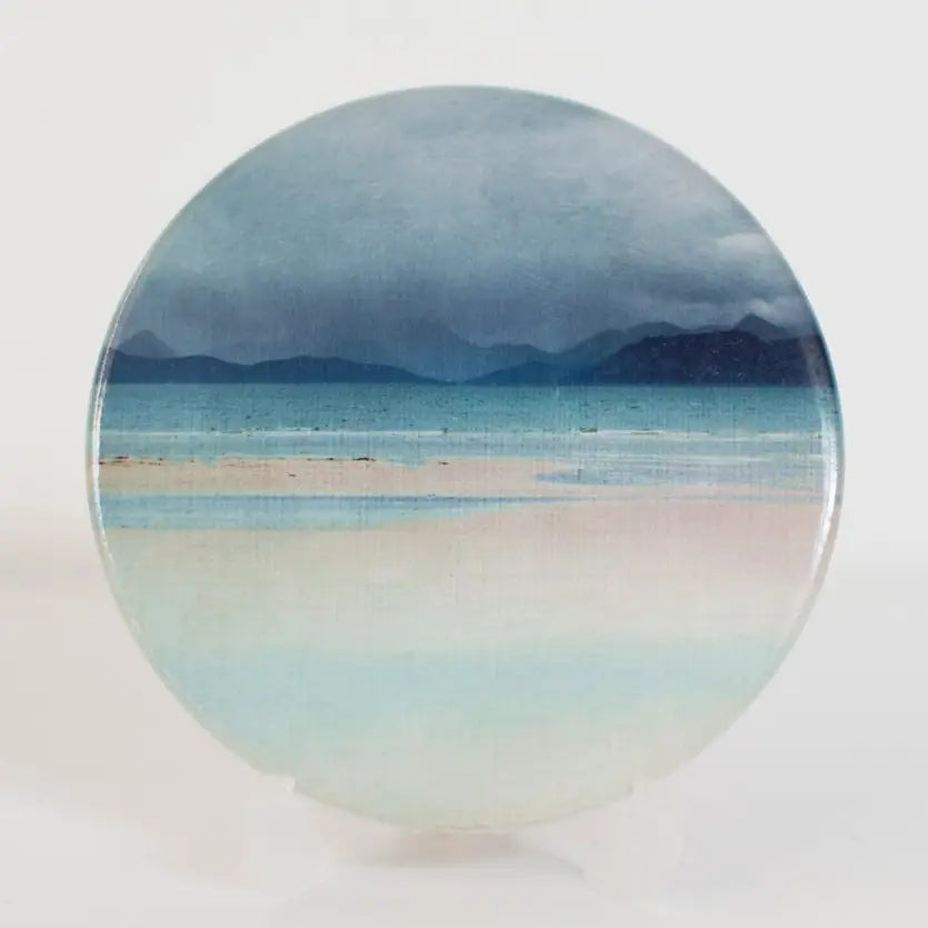 A round ceramic coaster featuring a scenic landscape print of a beach with mountains, displayed on a white surface against a white background.