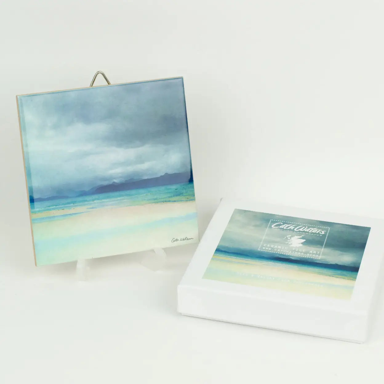 A square abstract beach landscape artwork shown standing on a small easel, with its white box lid placed beside it on a clean white background. The artwork features layered blue, gray, and pale aqua tones resembling sky, water, and sand.