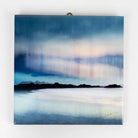 A square ceramic wall art tile print depicting a misty coastal landscape with dark mountain silhouettes and a soft blue, white, and pale pink sky, hanging on a plain white wall.