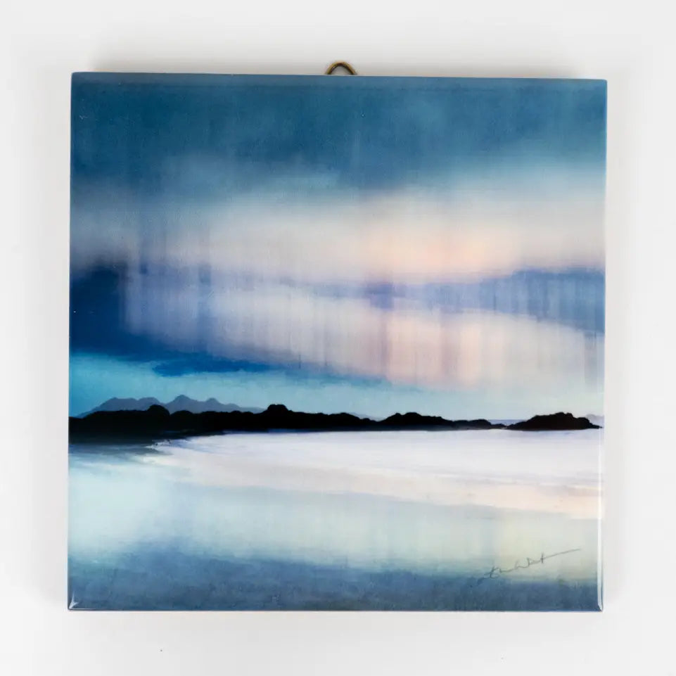 A square ceramic wall art tile print depicting a misty coastal landscape with dark mountain silhouettes and a soft blue, white, and pale pink sky, hanging on a plain white wall.