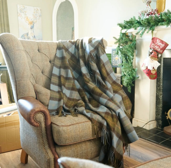 A beige upholstered wingback armchair in a decorated living room, with a large plaid throw blanket draped over the seat and back. The blanket has brown, beige, gray, and muted blue stripes with fringe detailing.