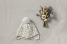 Knitted baby hat with a pom-pom and a small bouquet of flowers on a neutral background