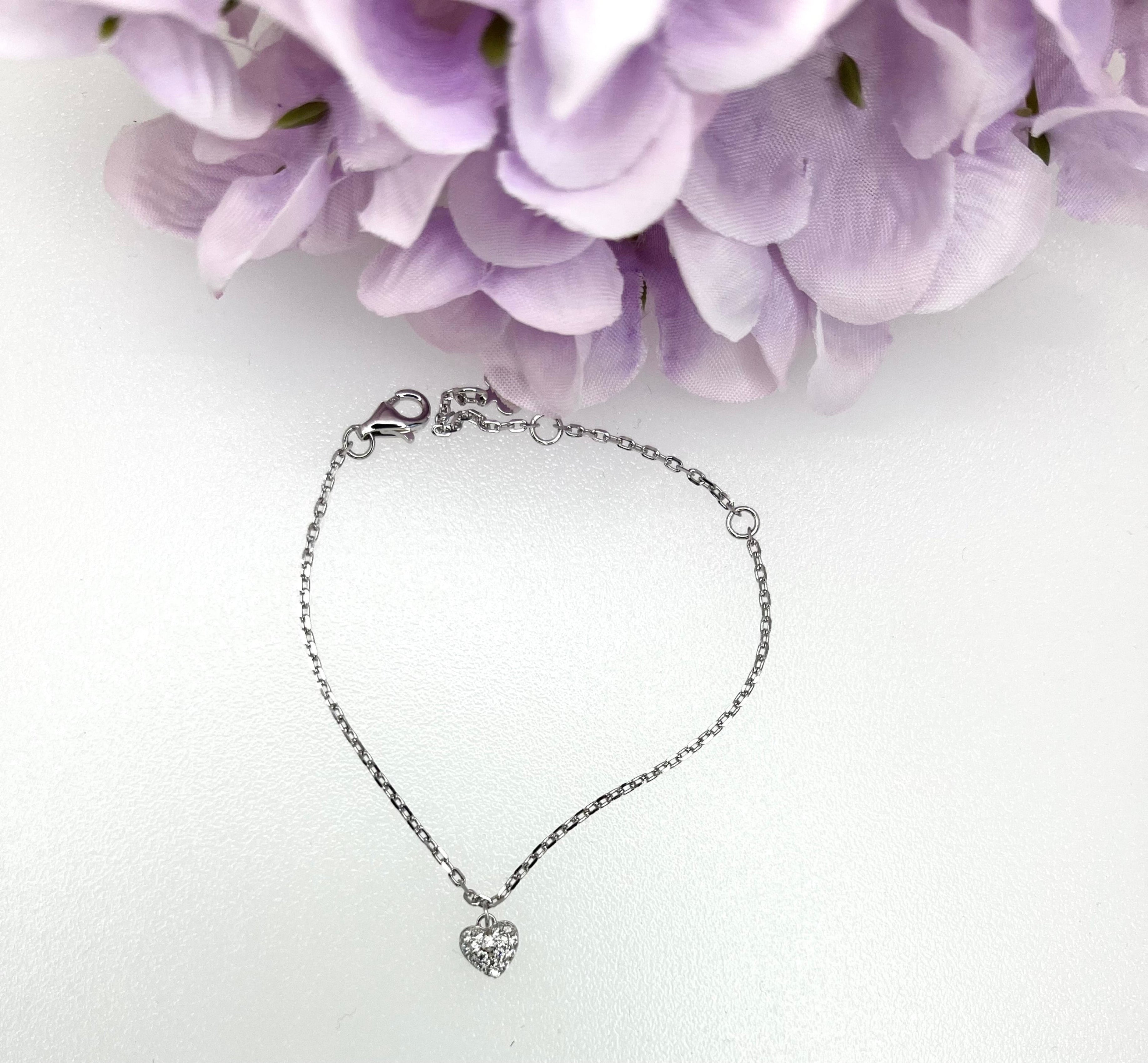 Silver bracelet with a heart-shaped charm on a white background with purple flowers.