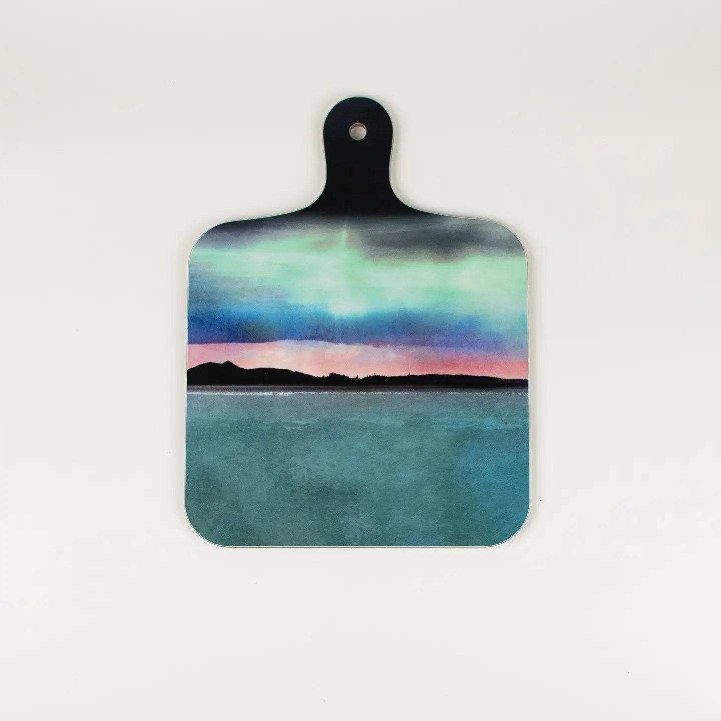 A square wooden cutting board with rounded corners and a black handle, featuring a printed landscape design of a teal sea under a colorful aurora sky, shown on a plain white background.