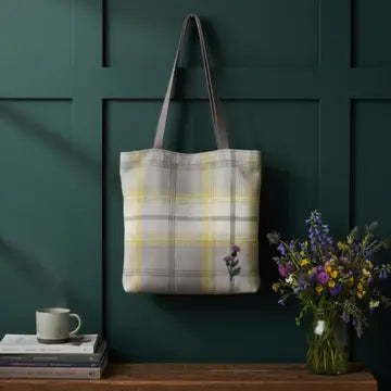 A plaid tote bag with gray handles hangs on a dark green paneled wall above a wooden shelf. The bag has a white, gray, and yellow plaid pattern with a small embroidered purple flower.