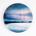 A round coaster featuring an abstract landscape painting of a blue and white seascape with a dark silhouette of mountains or land across the horizon, shown against a plain white background.