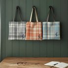 Three plaid fabric tote bags hang on a dark green paneled wall above a wooden table with an open notebook and glasses. The tote bags have dark brown or beige handles and small embroidered purple flowers near the bottom left corner.