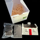 A DIY safe and house kit shown with its white cardboard box, a vacuum-sealed package of dark brown material, a flat gray sheet, and a small white house mold with a dark brown roof and red door, all set against a solid black background.