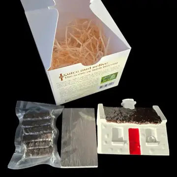 A DIY safe and house kit shown with its white cardboard box, a vacuum-sealed package of dark brown material, a flat gray sheet, and a small white house mold with a dark brown roof and red door, all set against a solid black background.