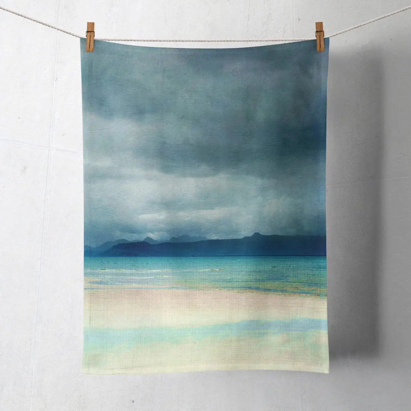 A rectangular textile print or towel hanging from a string with two wooden clips against a light gray wall. The printed scene shows a seascape with dark blue-gray stormy sky, turquoise water, distant dark mountains, and pale sandy shore.