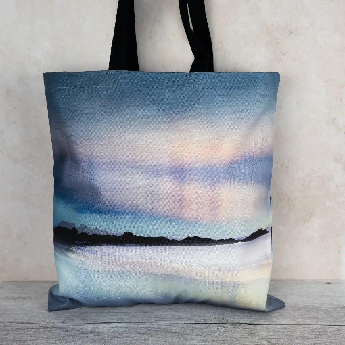 A canvas tote bag with black handles, featuring a printed landscape design of a beach and mountains under a blue and purple sky, set against a textured beige wall and gray wooden floor.