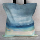 A canvas tote bag featuring a painted beach landscape design in shades of blue, teal, and cream, with black shoulder straps, resting on a rustic wooden surface against a neutral beige wall.