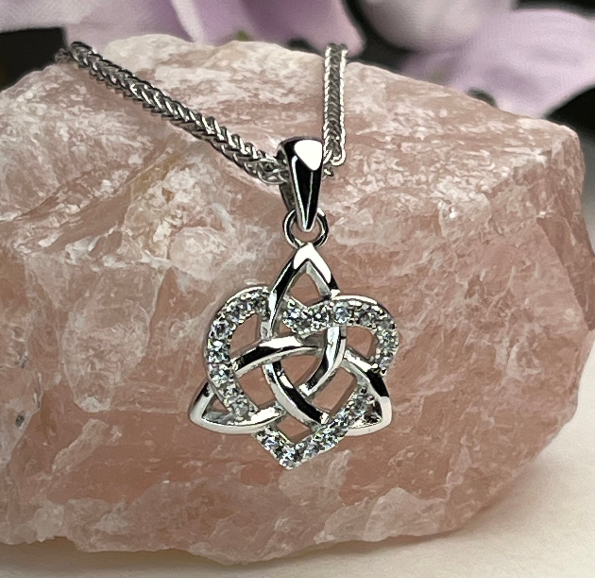 A silver Celtic knot triquetra pendant necklace with clear stones, resting on a pink rose quartz crystal.