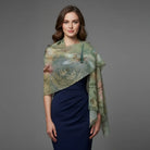 A woman modeling a sheer, semi-transparent shawl with a dragonfly and floral print in muted green, beige, and brown tones, draped over a navy blue dress against a solid gray studio background.