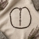 A dark brown beaded rosary necklace with a silver-toned crucifix pendant and a round medallion, arranged on a textured cream linen fabric with decorative stones and dried flowers.