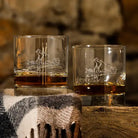 Two clear whiskey glasses with etched golf-themed designs, filled with amber liquor, set on a rustic wooden surface with a plaid blanket in the foreground and a blurred stone wall behind.
