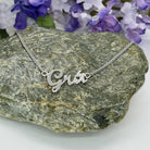 A silver script name necklace reading 'Grá' is draped over a textured gray stone, with purple and white flowers blurred in the background.