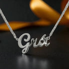A silver script nameplate necklace spelling 'Grá' on a silver chain, displayed against a dark background with a blurred gold ribbon.