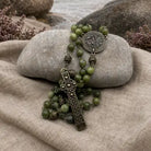 An olive green beaded rosary with an ornate bronze cross and medallion, draped over a smooth gray stone on a beige linen cloth with rocks in the background.