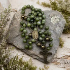 An olive green beaded rosary with antique bronze metal accents, including a central medallion and a cross, arranged on a gray stone surrounded by small white flowers and greenery.