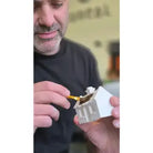 A man in a black t-shirt holds a small white miniature house model while applying gold-colored paint with a fine brush.