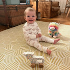Baby wearing highland coo sleeper sitting on a patterned rug with toys in a home setting