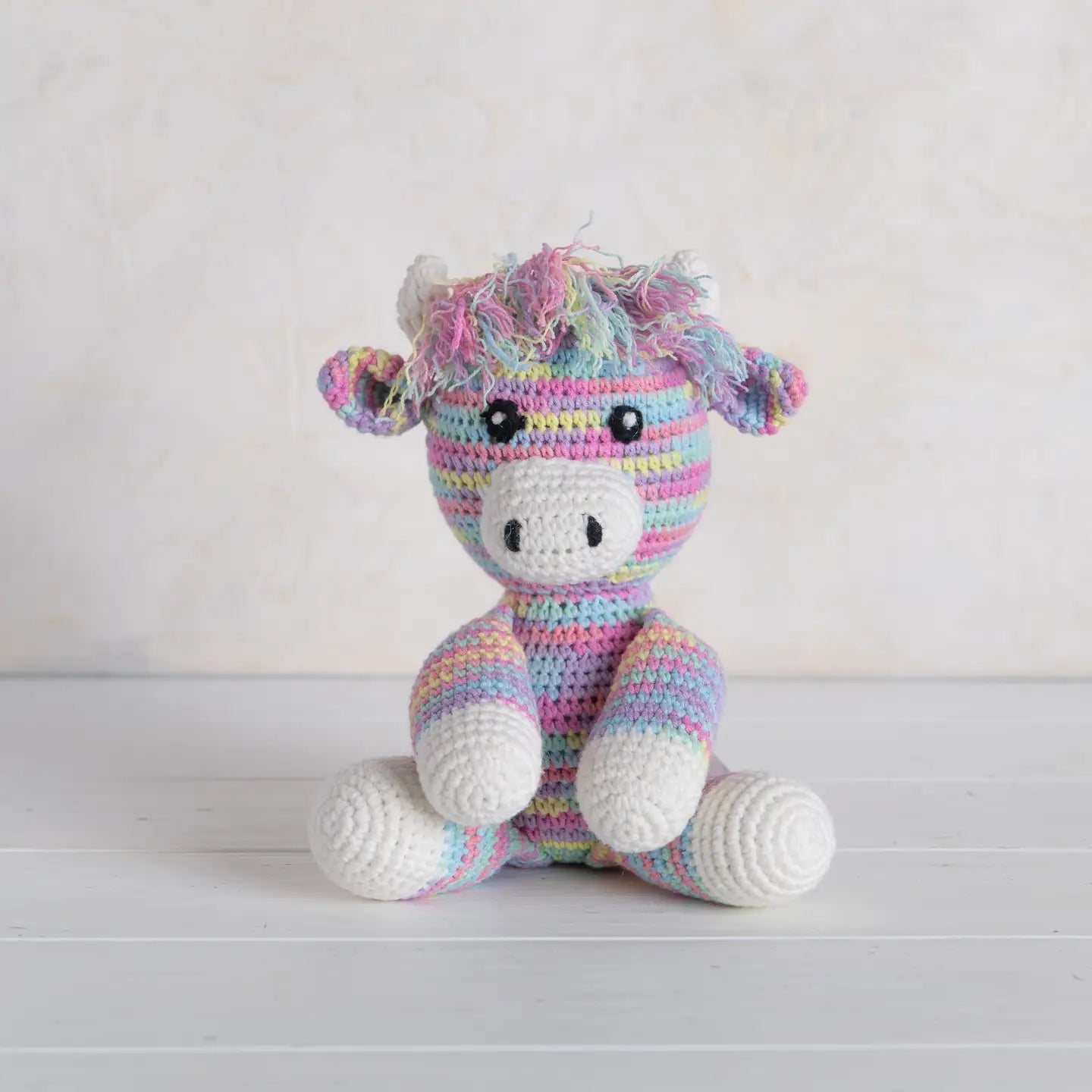 Crocheted highland cow toy with multicolored mane on a white wooden surface