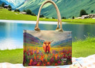 Tote bag with a scenic design of a cow in a field, placed on grass with a lake and mountains in the background.