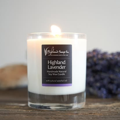 A lit Highland Lavender soy wax candle in a clear glass jar, set on a rustic wooden surface with blurred lavender buds in the background.