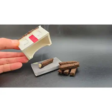 A hand holds a miniature off-white plastic house with a red window detail, next to several brown textured log pieces on a gray surface.